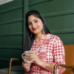 Positive smiling Indian lady in stylish traditional wear sitting on chair against green wall with cup of hot drink and looking at camera