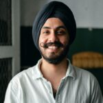 Positive bearded Indian male in shirt and turban standing in room and smiling while looking at camera